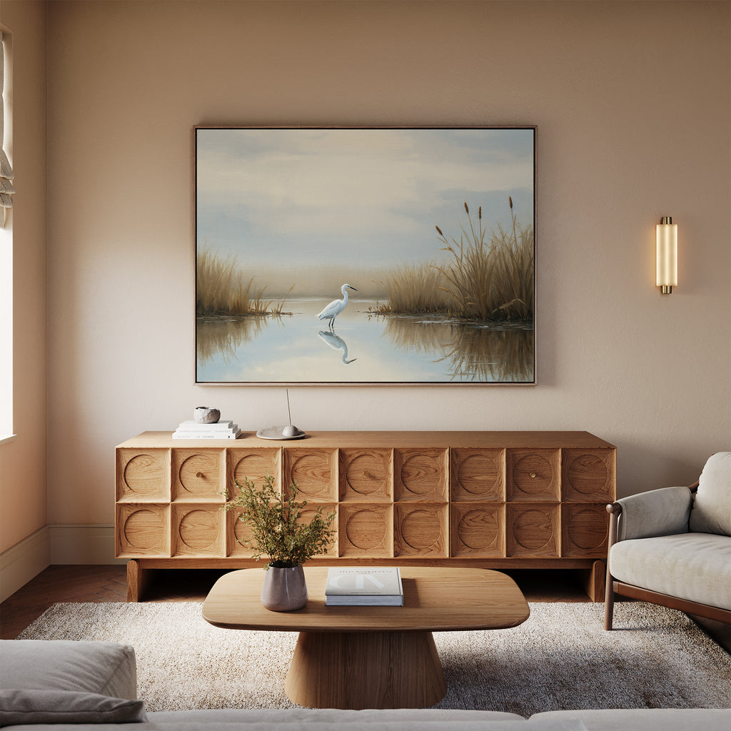 A cozy living room with a modern wooden cabinet, coffee table, light sofa, wall-mounted sconce, and serene decor—a large Heron's Morning Canvas Art - Horizontal above the cabinet showing a calm pond with reeds.