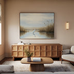A cozy living room with a modern wooden cabinet, coffee table, light sofa, wall-mounted sconce, and serene decor—a large Heron's Morning Canvas Art - Horizontal above the cabinet showing a calm pond with reeds.
