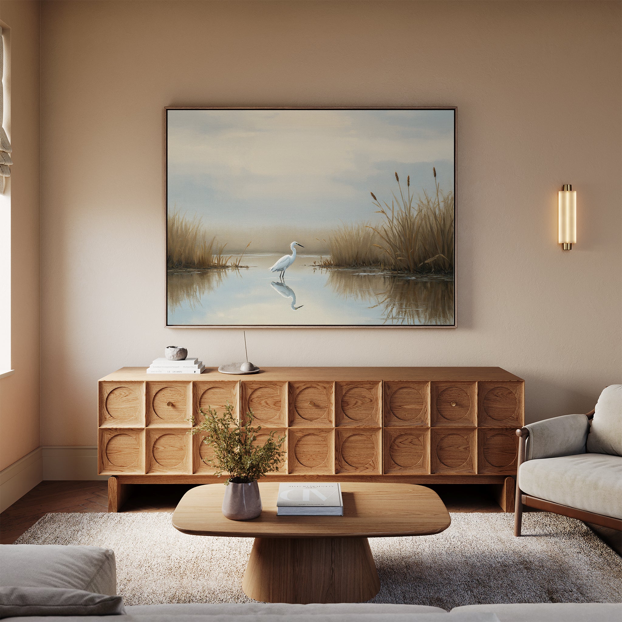 A cozy living room with a modern wooden cabinet, coffee table, light sofa, wall-mounted sconce, and serene decor—a large Heron's Morning Canvas Art - Horizontal above the cabinet showing a calm pond with reeds.