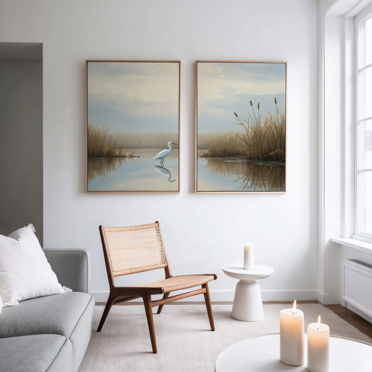 A modern living room with a gray sofa, woven chair, round white table, and lit candles. Above the chair hang "Heron's Morning - Set Of 2" art prints, featuring serene wetland scenes. Sunlight pours in through a large window.