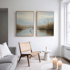 A modern living room with a gray sofa, woven chair, round white table, and lit candles. Above the chair hang "Heron's Morning - Set Of 2" art prints, featuring serene wetland scenes. Sunlight pours in through a large window.