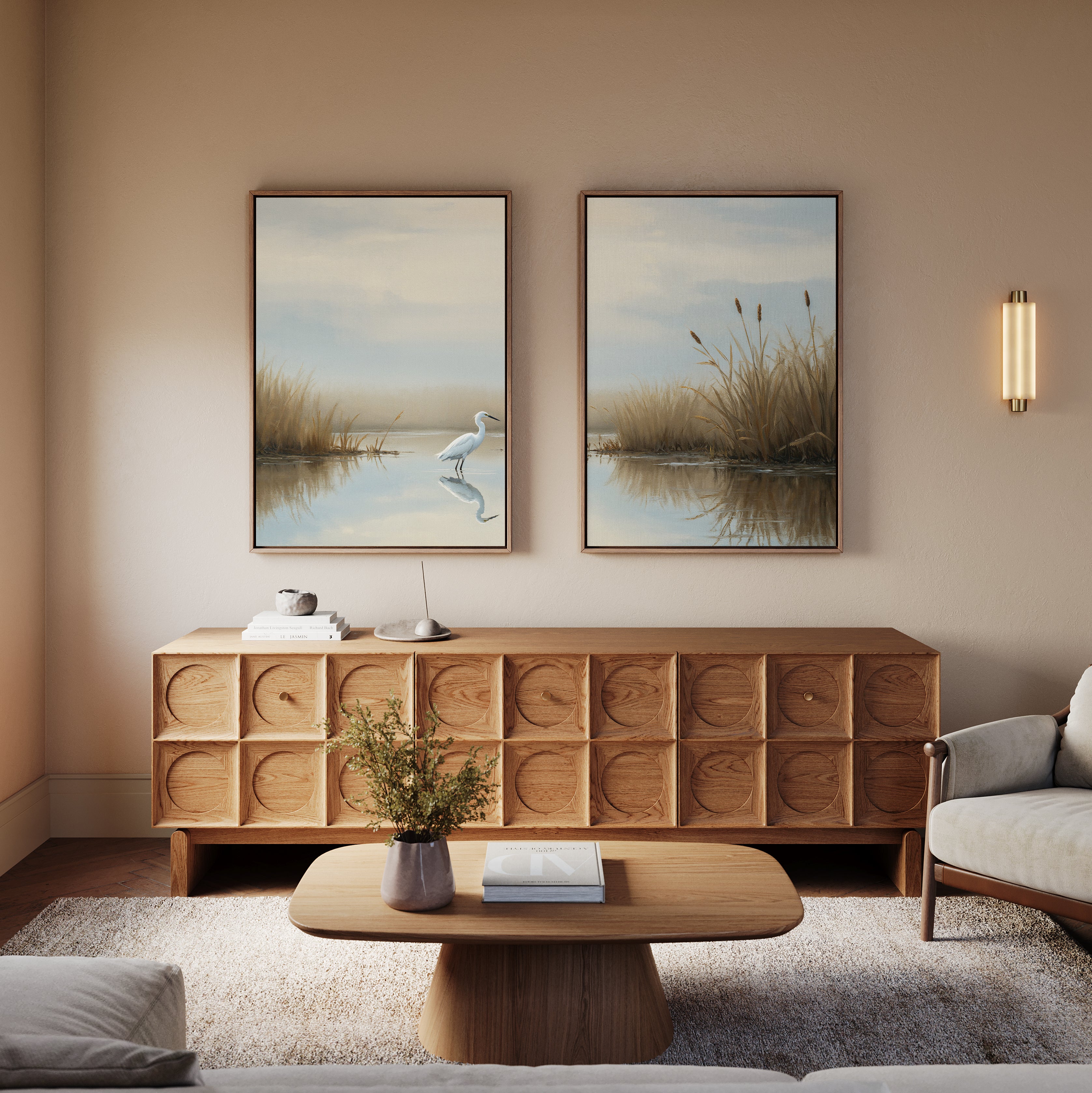 A cozy living room features a wooden sideboard and coffee table, a light sofa, and "Heron's Morning - Set Of 2" wall art on the beige wall. A small plant and books decorate the tables.