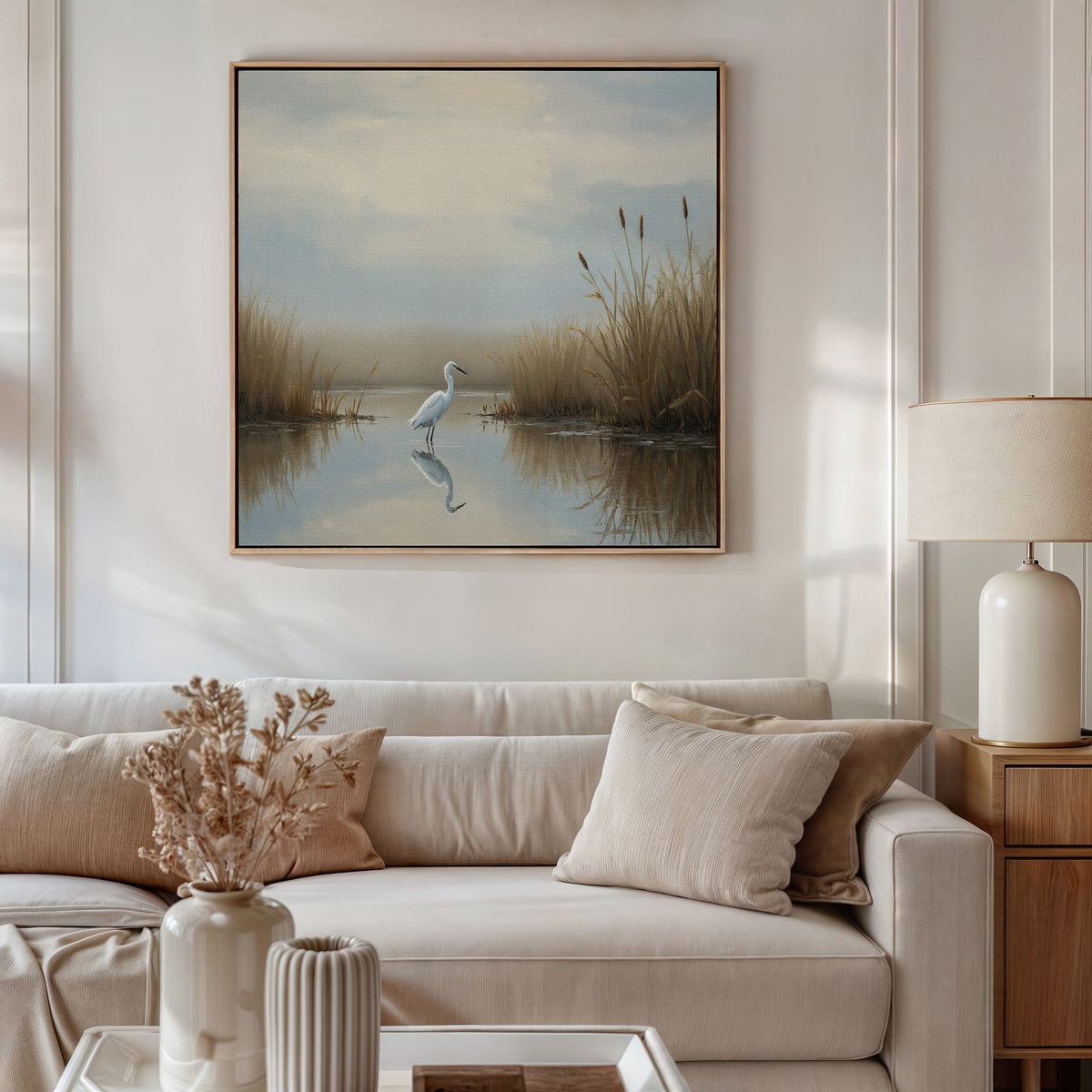 A cozy living room features a white sofa with beige pillows, a table with dried flowers, and a lamp. Above the sofa hangs the Heron's Morning Canvas Art - Square, depicting a white egret in a tranquil wetland under cloudy skies.