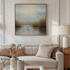 A cozy living room features a white sofa with beige pillows, a table with dried flowers, and a lamp. Above the sofa hangs the Heron's Morning Canvas Art - Square, depicting a white egret in a tranquil wetland under cloudy skies.