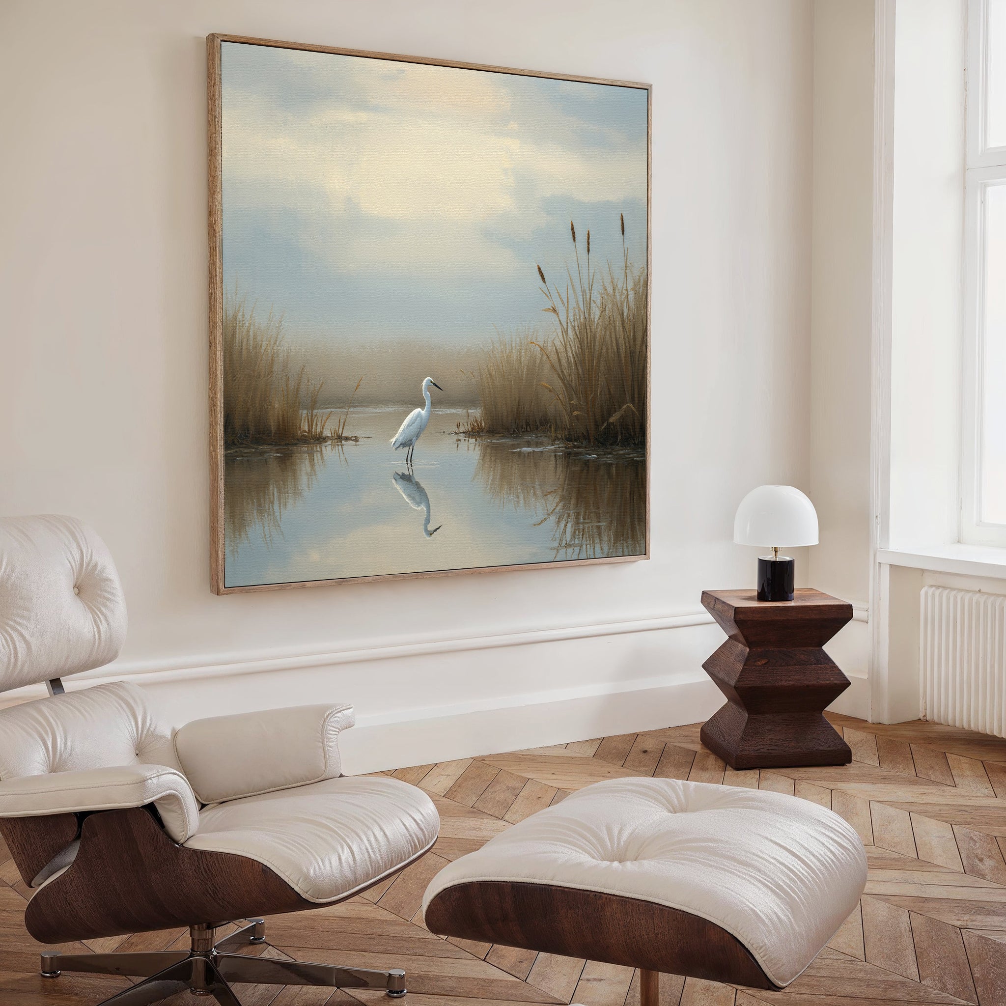 A modern living room with a white leather lounge chair and ottoman, a geometric side table and lamp, and Heron's Morning Canvas Art - Square depicting a serene egret scene displayed above the sofa.