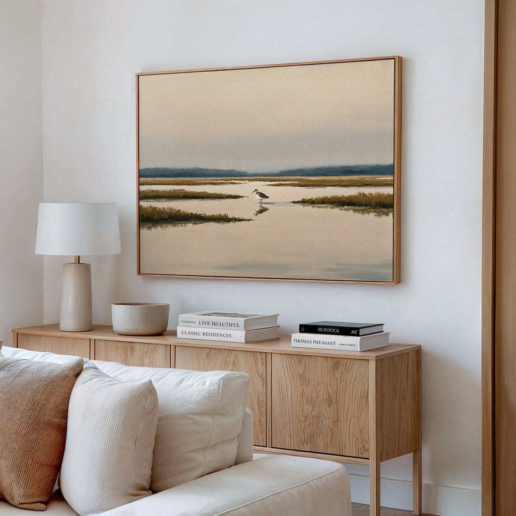 A modern living room features a light wood sideboard with books, a ceramic bowl, and a white table lamp. Above hangs the Marsh Bird Canvas Art - Horizontal, depicting a tranquil marsh scene. A cream sofa sits in the foreground.