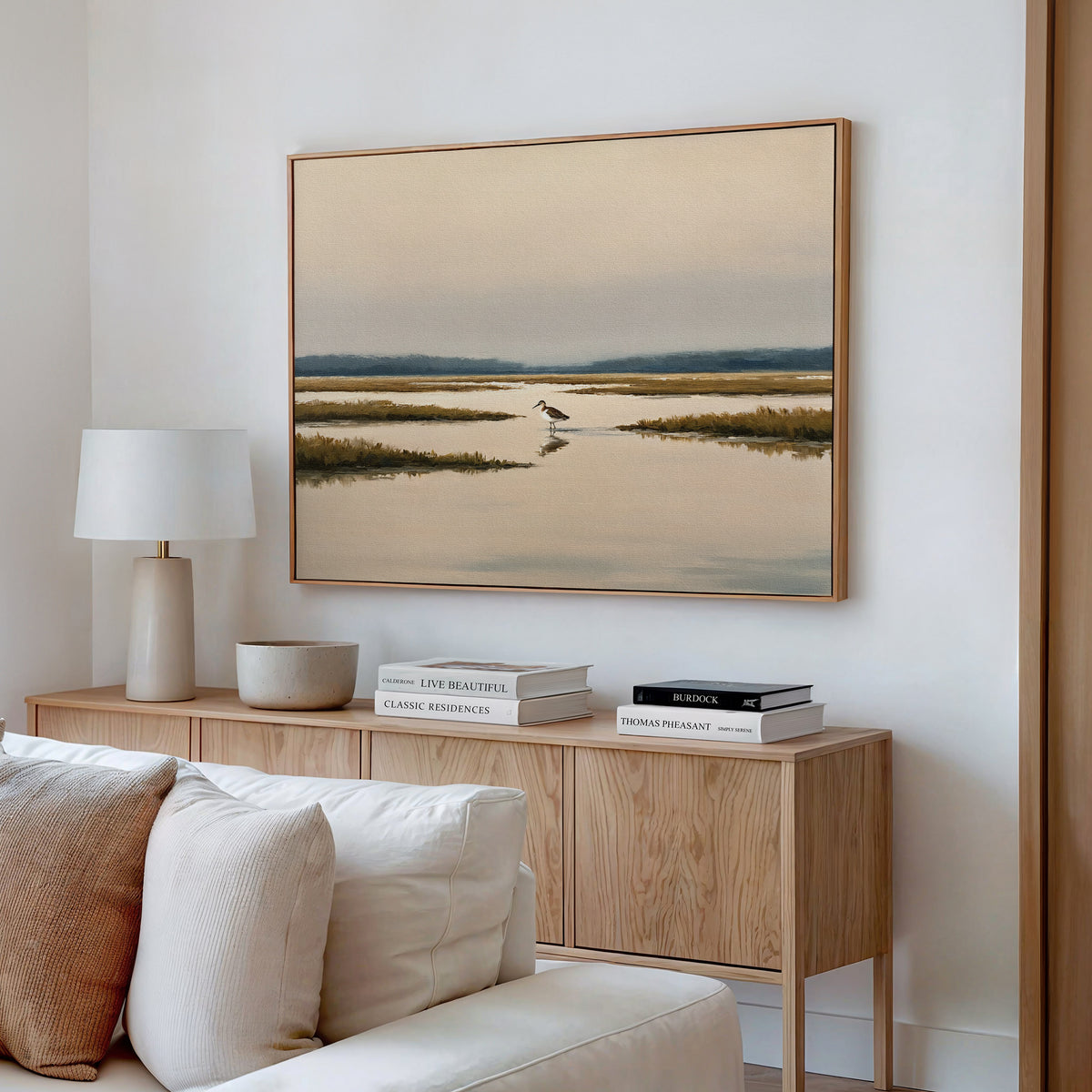 A modern living room features a light wood sideboard with books, a ceramic bowl, and a white table lamp. Above hangs the Marsh Bird Canvas Art - Horizontal, depicting a tranquil marsh scene. A cream sofa sits in the foreground.