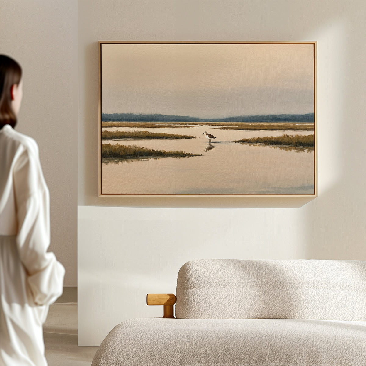A person stands in a softly lit room, admiring the Marsh Bird Canvas Art - Horizontal—a large landscape of calm water and marsh grasses—framed above a beige couch.