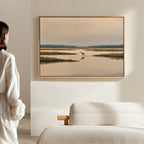 A person stands in a softly lit room, admiring the Marsh Bird Canvas Art - Horizontal—a large landscape of calm water and marsh grasses—framed above a beige couch.