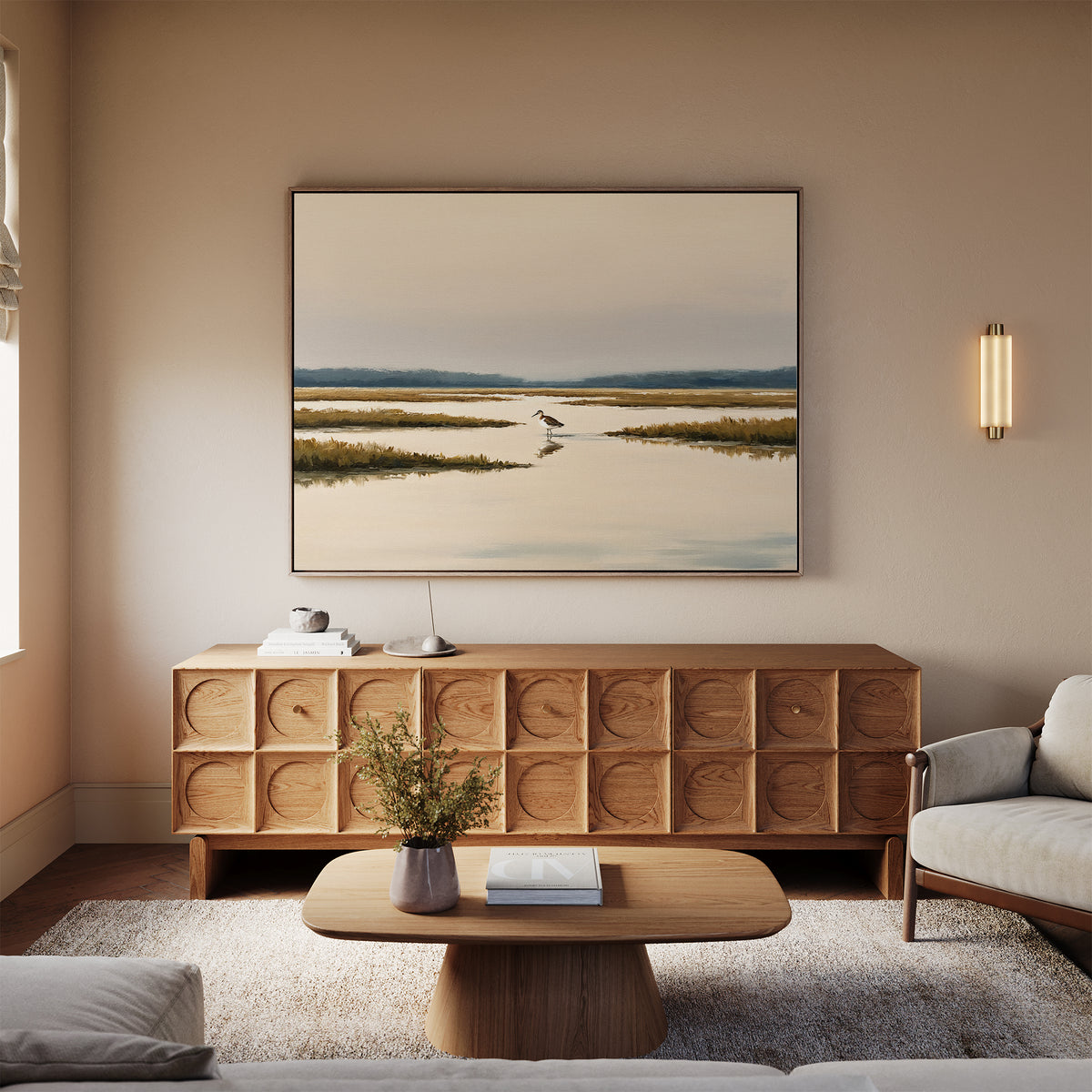 A cozy living room with a beige sofa, wooden coffee table, and a patterned sideboard. Above the sideboard hangs the Marsh Bird Canvas Art - Horizontal, illuminated by a sconce light on the wall.
