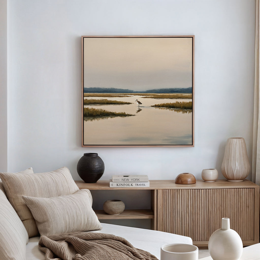 A modern living room features a beige sofa, neutral cushions, a wooden sideboard with vases and books, and above it hangs the Marsh Bird Canvas Art - Square, a tranquil framed landscape for a serene touch.