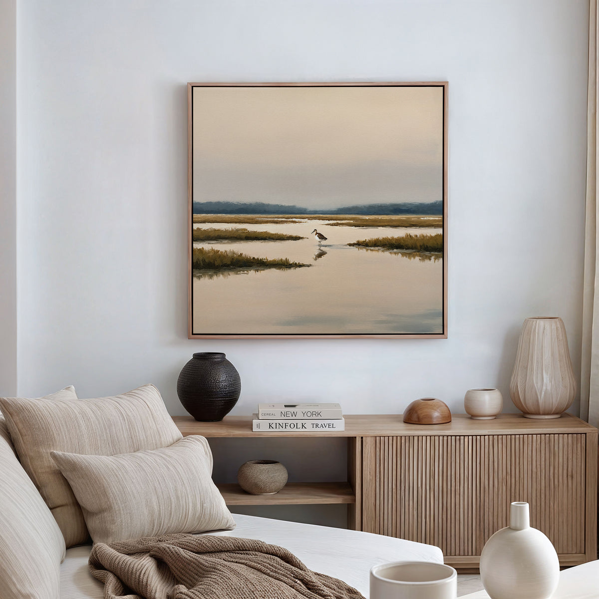 A modern living room features a beige sofa, neutral cushions, a wooden sideboard with vases and books, and above it hangs the Marsh Bird Canvas Art - Square, a tranquil framed landscape for a serene touch.