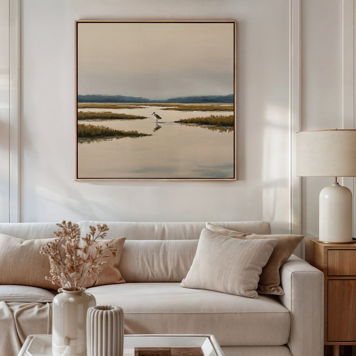 A modern living room with a beige sofa, neutral pillows, a vase of dried flowers, a table lamp, and tranquil wall art—a framed Marsh Bird Canvas Art - Square—hanging above the sofa.