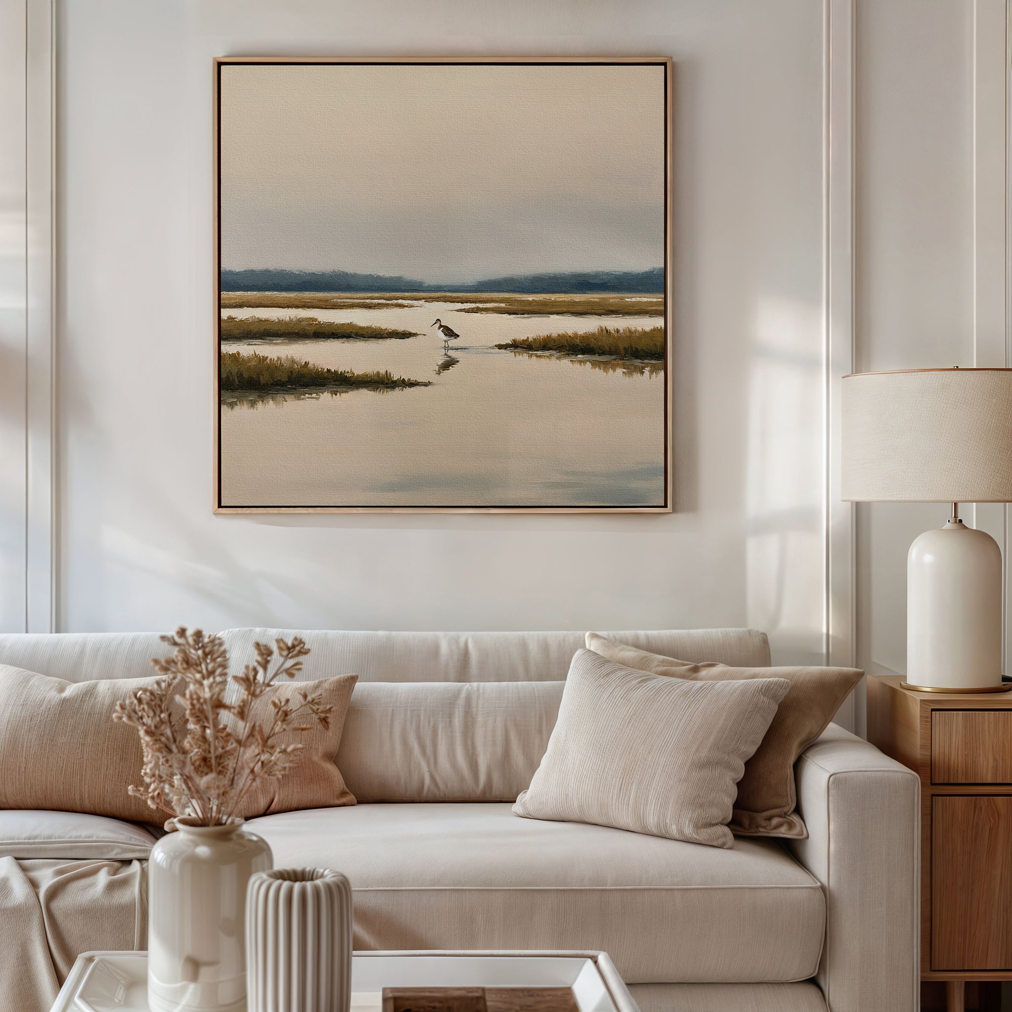 A modern living room with a beige sofa, neutral pillows, a vase of dried flowers, a table lamp, and tranquil wall art—a framed Marsh Bird Canvas Art - Square—hanging above the sofa.