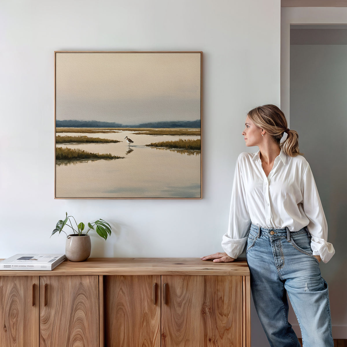A woman in a white shirt and jeans stands by a wooden cabinet, admiring the Marsh Bird Canvas Art - Square—a serene marshland print with water, grass, and a distant bird—hanging on the white wall above.