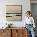 A woman in a white shirt and jeans stands by a wooden cabinet, admiring the Marsh Bird Canvas Art - Square—a serene marshland print with water, grass, and a distant bird—hanging on the white wall above.