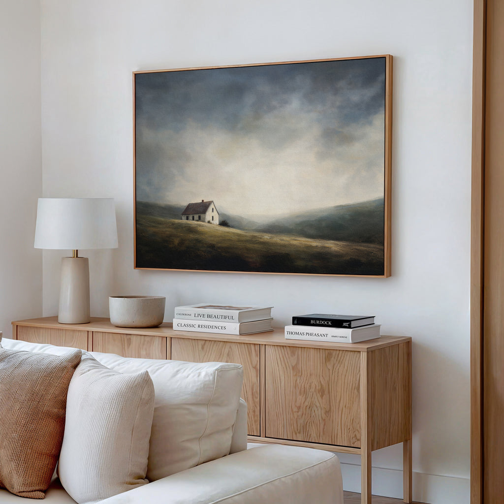 A cozy living room has a wooden sideboard with books, a lamp, and a bowl. Above it hangs the Highland Solitude Canvas Art - Horizontal, its moody landscape adding rustic tranquility above the cream-colored sofa.