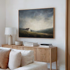 A cozy living room has a wooden sideboard with books, a lamp, and a bowl. Above it hangs the Highland Solitude Canvas Art - Horizontal, its moody landscape adding rustic tranquility above the cream-colored sofa.