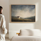A woman in white stands in a bright, minimal room, facing the Highland Solitude Canvas Art - Horizontal—a moody landscape of a lone house on a grassy hill under cloudy skies—displayed above a beige sofa.