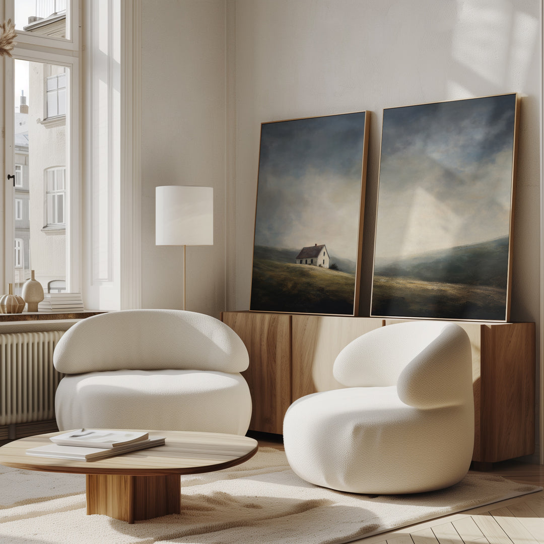 A modern living room with two cushioned white chairs, a round wooden coffee table, a wooden sideboard, and the Highland Solitude - Set of 2 moody landscape canvases leaning beneath a tall window bathed in natural light.