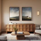 A cozy living room features a wooden sideboard with geometric patterns, a matching coffee table, a light gray sofa, and two framed moody landscape paintings above—the Highland Solitude - Set of 2.