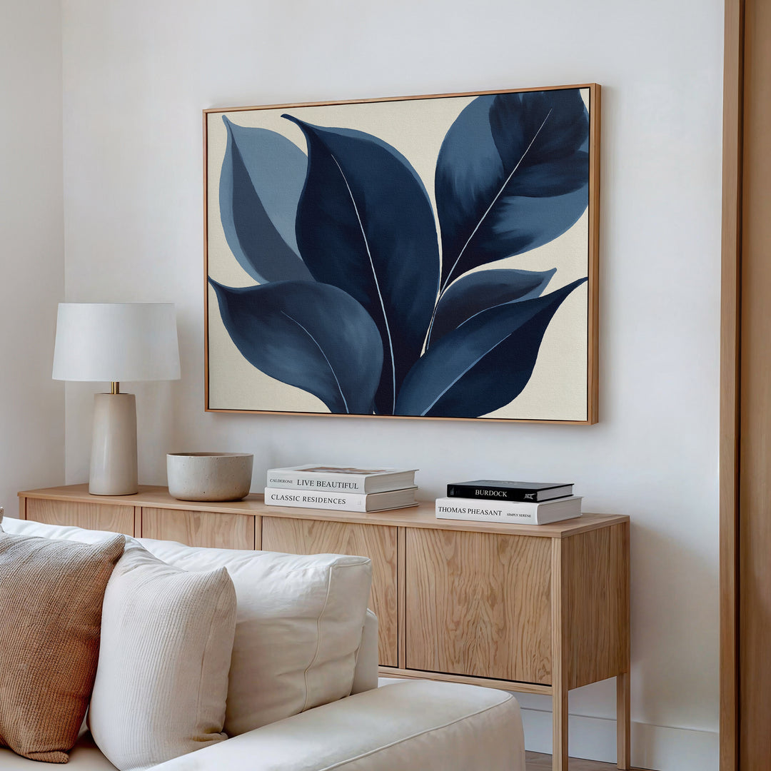 A modern living room features a wooden sideboard with books, a lamp, and ceramics, topped by the Indigo Depths Canvas Art - Horizontal—a navy botanical piece—displayed on a beige wall above a light-colored sofa.