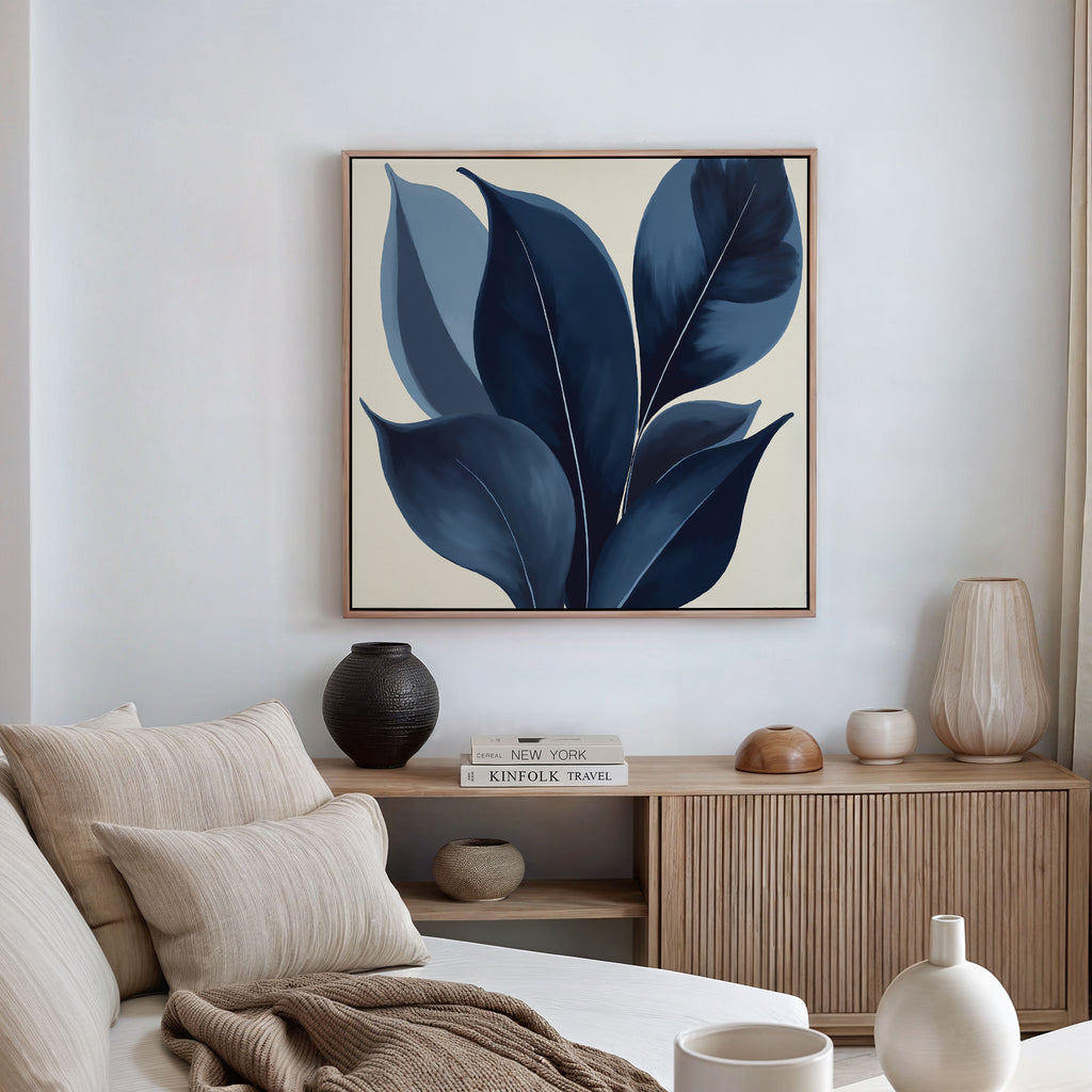 A modern living room features a beige couch, neutral cushions, and a wooden sideboard with ceramic vases, books, and a bowl. Above the sideboard hangs the Indigo Depths Canvas Art - Square, bringing elegant navy botanical style to the space.