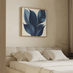A minimalist bedroom features a beige upholstered bed with white bedding and four pillows. Above the bed hangs the Indigo Depths Canvas Art - Square, showcasing large blue leaves. A bare branch in a vase sits on the side table.