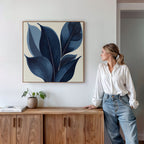 A woman in a white blouse and jeans leans against a wooden cabinet, admiring the Indigo Depths Canvas Art - Square, a striking navy botanical piece on the wall. A small potted plant sits atop the cabinet.