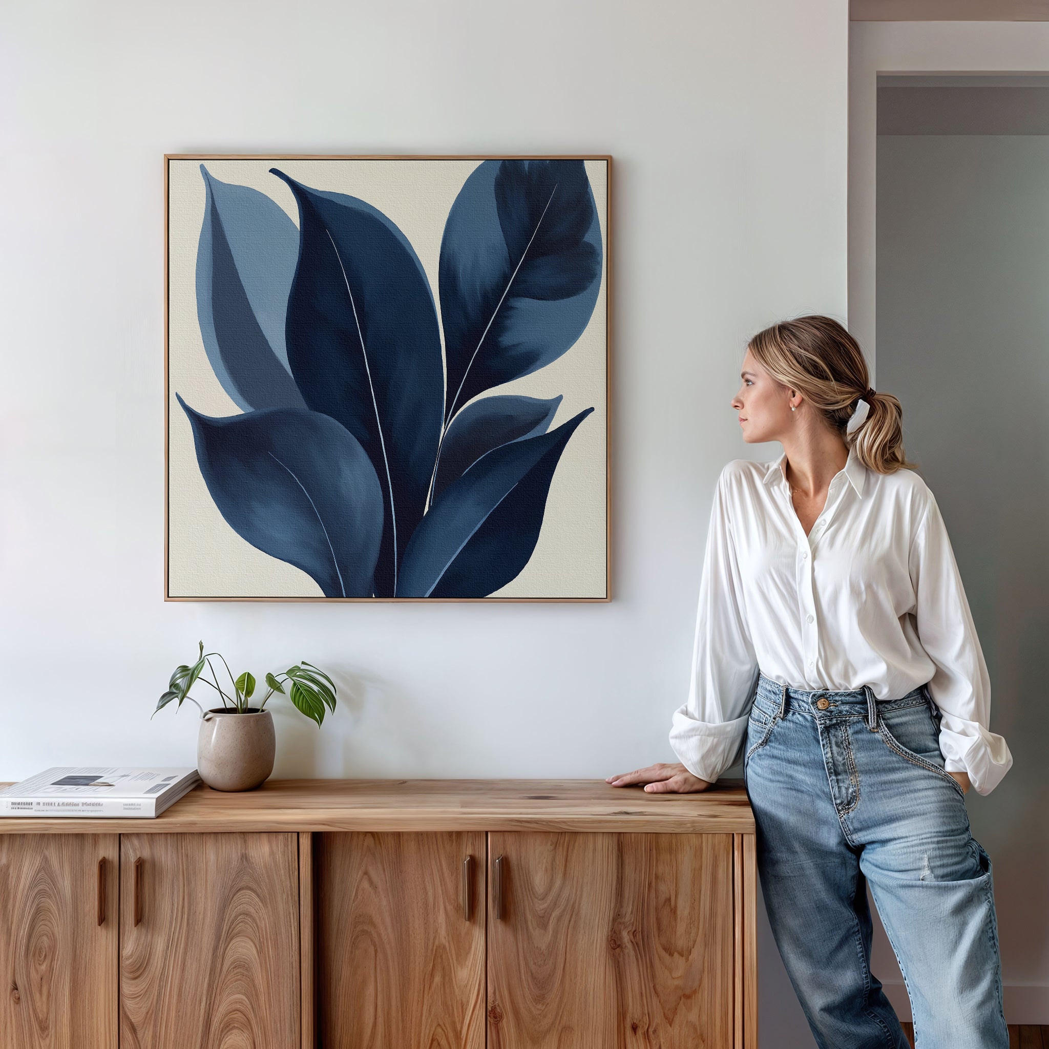 A woman in a white blouse and jeans leans against a wooden cabinet, admiring the Indigo Depths Canvas Art - Square, a striking navy botanical piece on the wall. A small potted plant sits atop the cabinet.