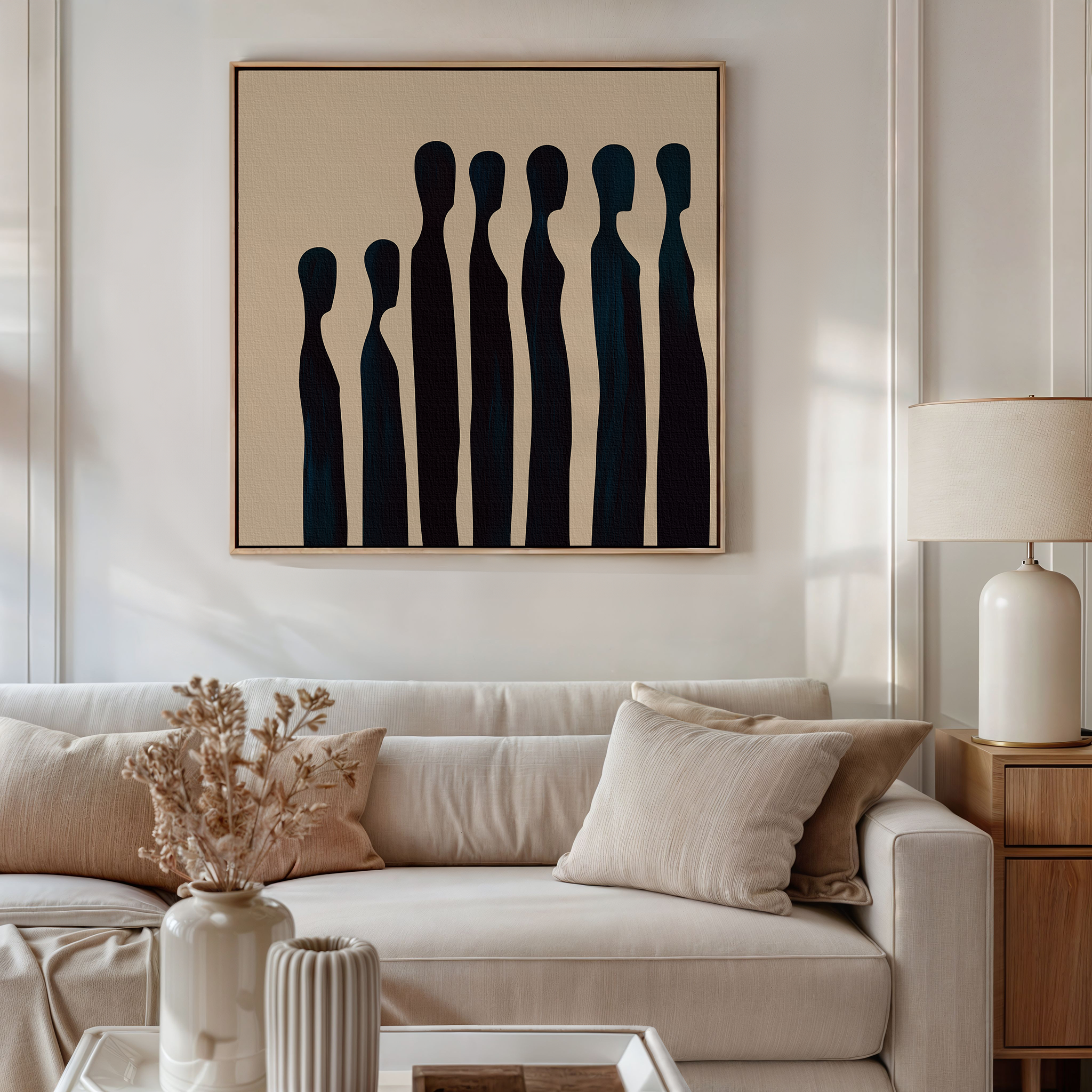 A cozy living room features a beige sofa, neutral pillows, a wooden side table with a white lamp, dried flowers in a vase, and the Indigo Profile Abstract Canvas Art with tall black shapes on a beige background hanging above the sofa.