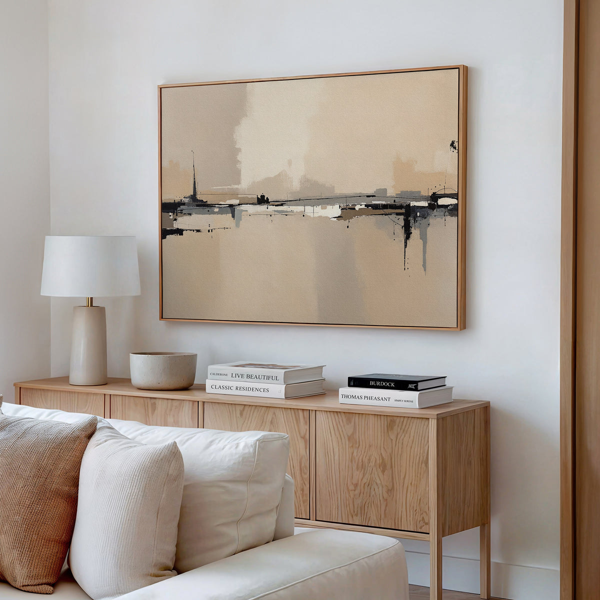 A minimalist living room with a light wood sideboard, stacked books, a white lamp, a bowl, and the Industrial Cityscape Canvas Art - Horizontal above a white sofa with textured pillows.