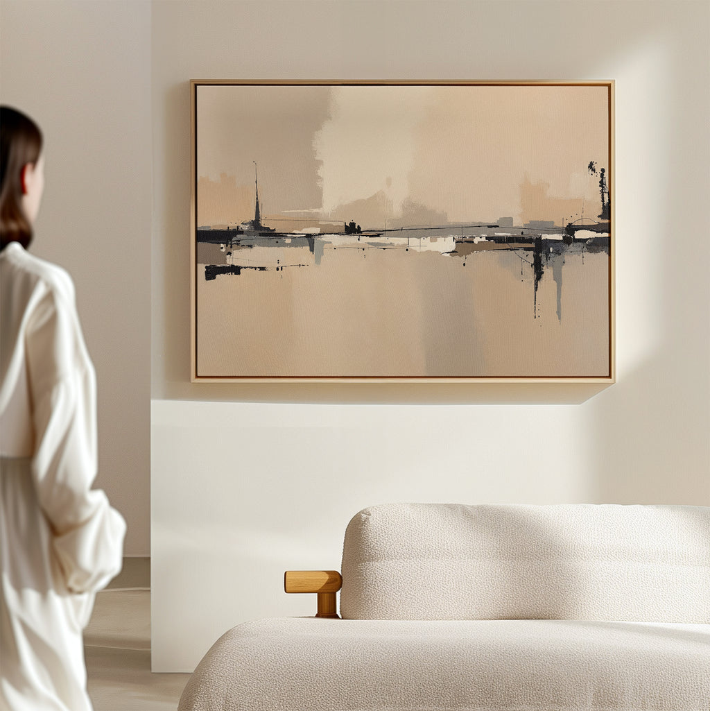 A woman in a white outfit stands in a minimalist room, admiring the Industrial Cityscape Canvas Art - Horizontal with beige, black, and gray tones hanging above a light-colored sofa.