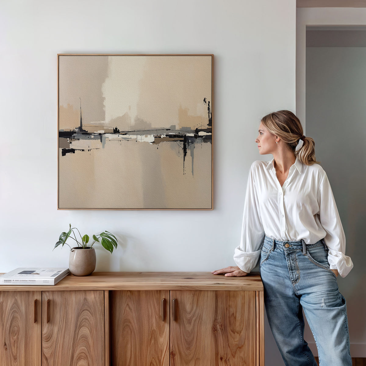 A woman in a white blouse and jeans leans against a wooden cabinet, gazing at the Industrial Cityscape Canvas Art - Square displayed above. A small potted plant and books on the cabinet complete this modern living room decor.