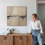 A woman in a white blouse and jeans leans against a wooden cabinet, gazing at the Industrial Cityscape Canvas Art - Square displayed above. A small potted plant and books on the cabinet complete this modern living room decor.