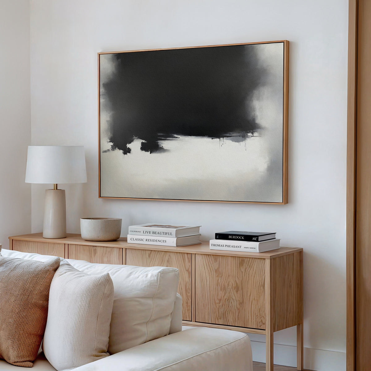 A modern living room features a light wooden sideboard with stacked books, a white table lamp, beige bowl, and the Ink Cloud Formation Canvas Art - Horizontal above a cream sofa with matching cushions for a sophisticated look.