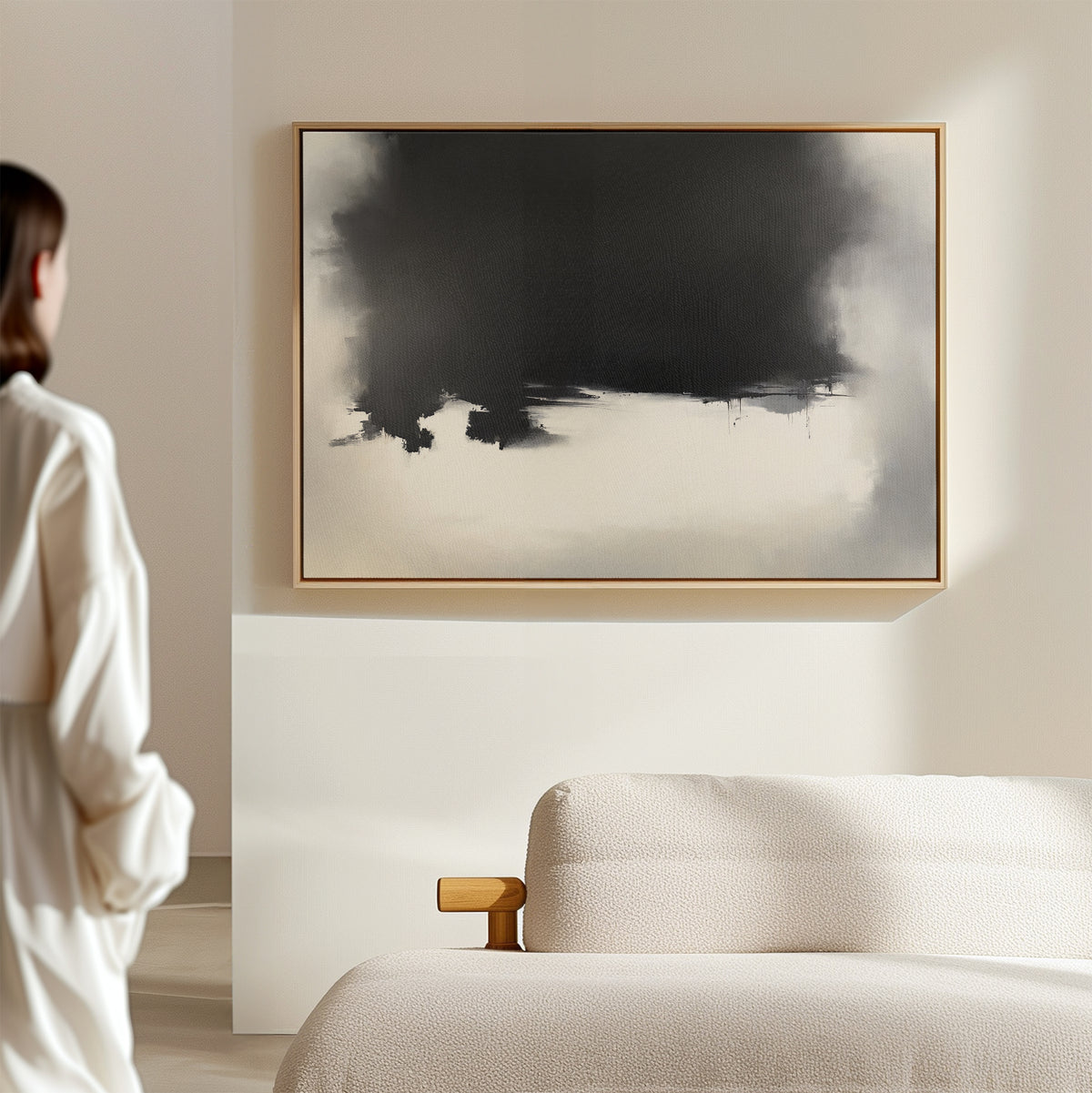 In a bright, minimal room, someone admires the Ink Cloud Formation Canvas Art - Horizontal, featuring bold black and white shapes above a beige couch and adding modern sophistication.