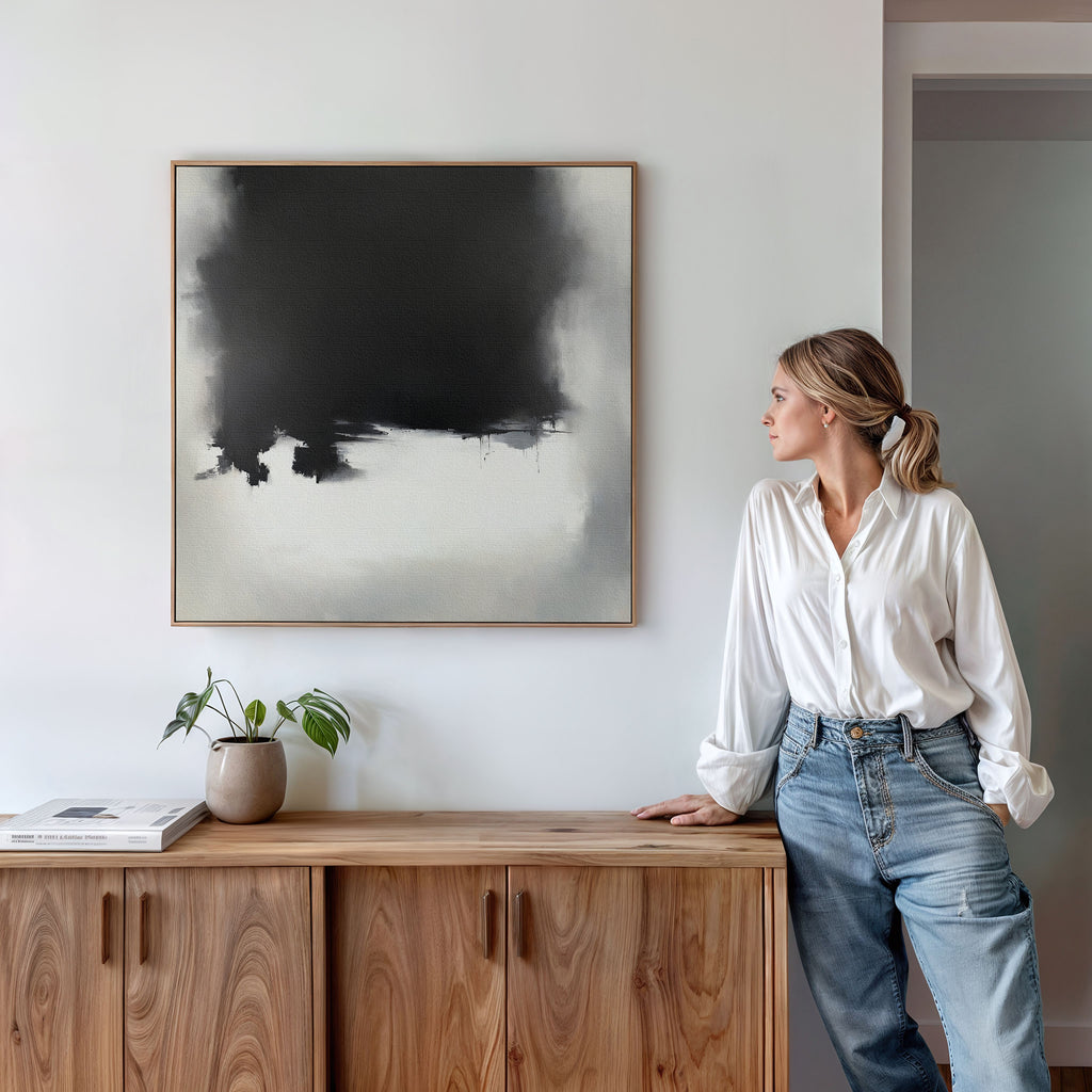 A woman in a white shirt and jeans leans on a wooden sideboard, admiring the Ink Cloud Formation Canvas Art - Square on the wall. A small plant and books beside her enhance the modern sophistication of this minimalist space.