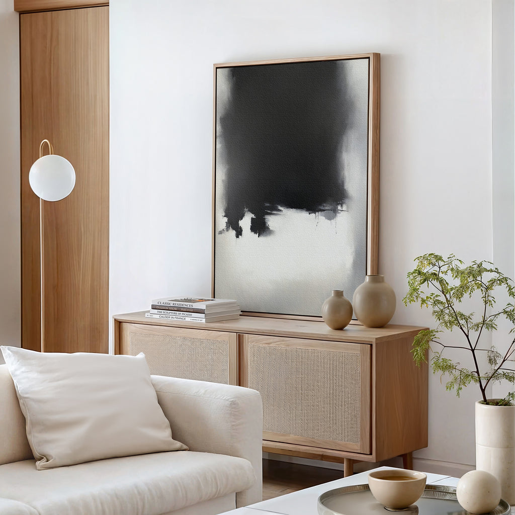 A modern living room with a light wood sideboard, Ink Cloud Formation Canvas Art - Vertical as a monochrome print, neutral vases, books, a round white lamp, a white sofa with cushions, and a small green potted plant for modern sophistication.