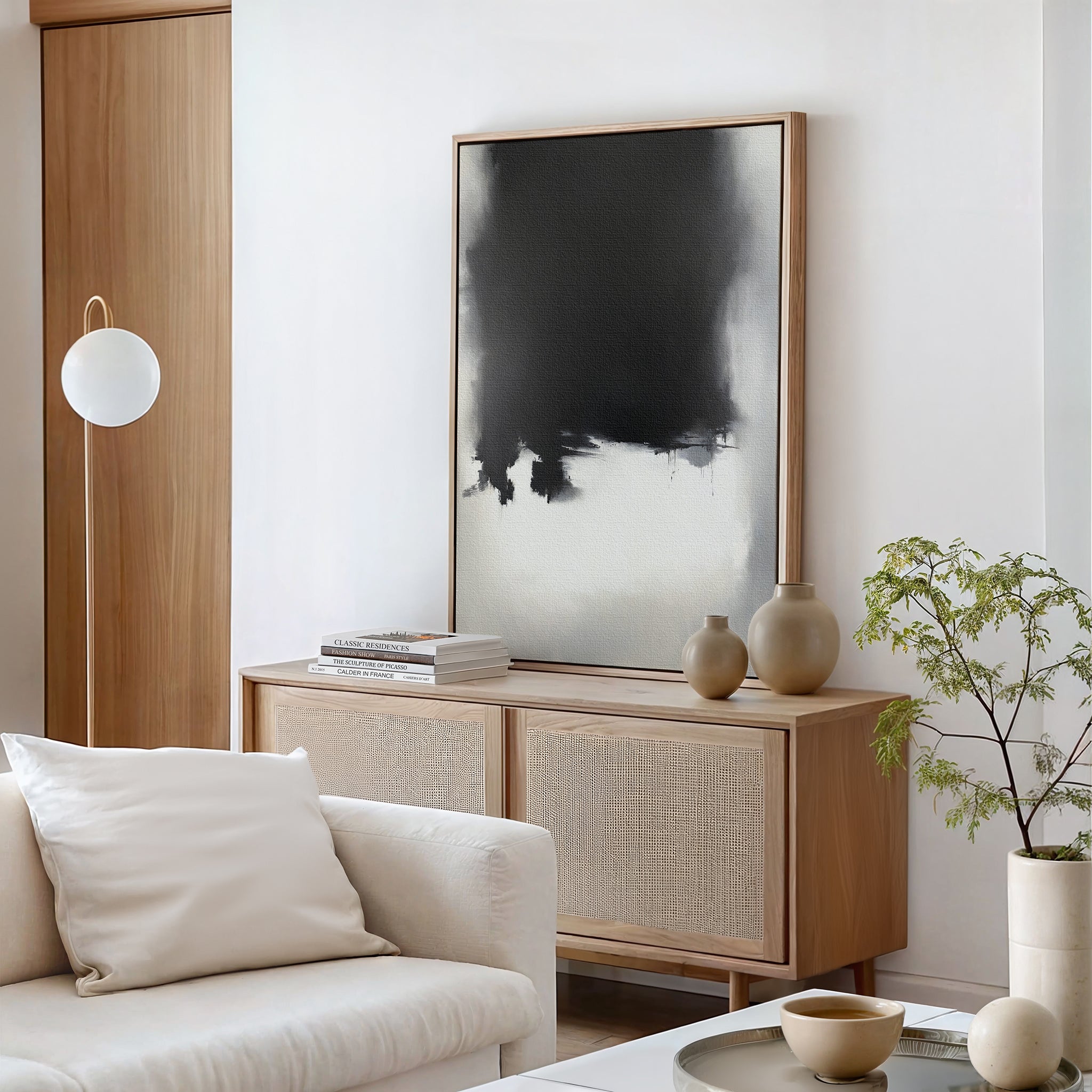 A modern living room with a light wood sideboard, Ink Cloud Formation Canvas Art - Vertical as a monochrome print, neutral vases, books, a round white lamp, a white sofa with cushions, and a small green potted plant for modern sophistication.