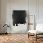 A minimal living room features a white cushioned chair, a round black side table with candles and books, and the Ink Cloud Formation Canvas Art - Vertical leaning against a white paneled wall with parquet wood flooring.