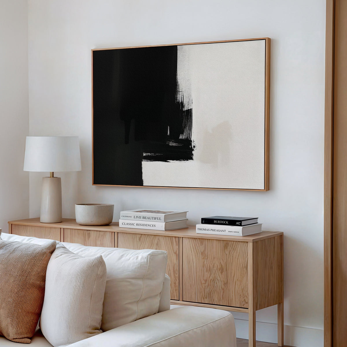 Minimalist living room with a light wooden console, stacked books, a white lamp, a bowl, and the Ink Divide Canvas Art - Horizontal (a large abstract black-and-white painting) on a white wall above a cream-colored sofa.