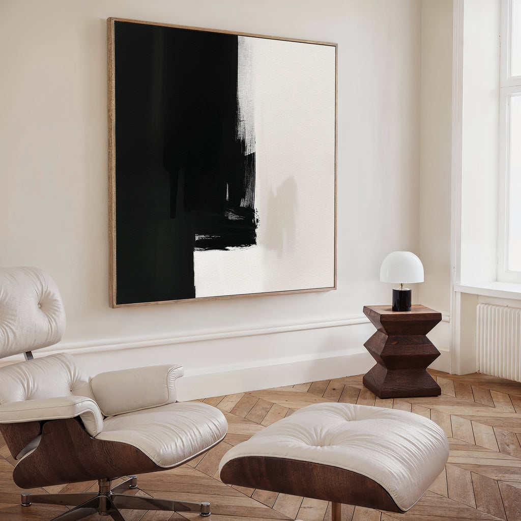 A modern, minimalist living room with a white leather lounge chair and ottoman, the Ink Divide Canvas Art - Square on the wall, a small wooden side table, and a round white lamp by a large window.
