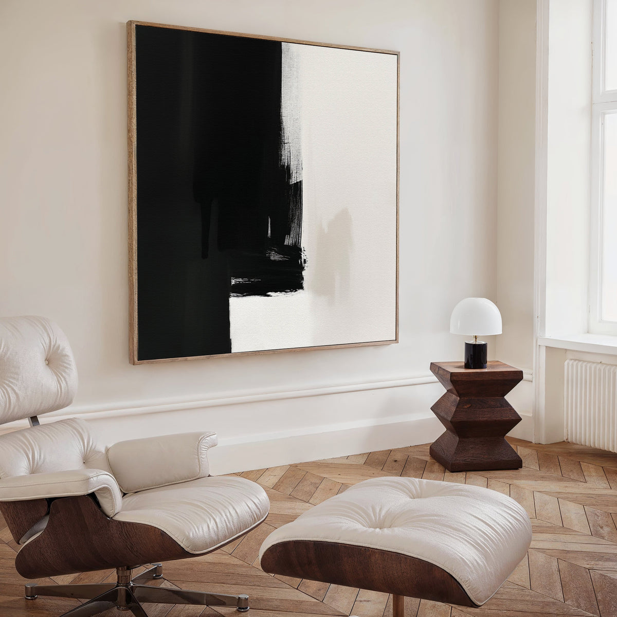 A modern, minimalist living room with a white leather lounge chair and ottoman, the Ink Divide Canvas Art - Square on the wall, a small wooden side table, and a round white lamp by a large window.