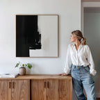 A woman in a white blouse and jeans leans on a wooden cabinet, gazing at the Ink Divide Canvas Art - Square above her. A potted plant and books on the cabinet enhance the serene, modern vibe of the space.