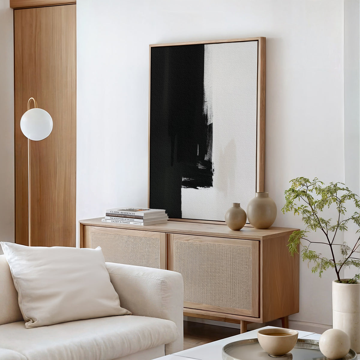 Minimalist living room featuring the Ink Divide Canvas Art - Vertical, a light wood cabinet, neutral pottery, a small potted plant, a white sofa with cushions, and a modern floor lamp in the corner.