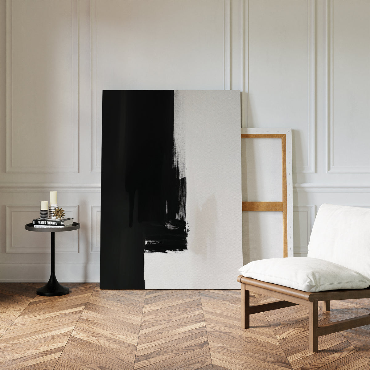 A minimalist room with a white cushioned chair, a round black side table with candles and books, and the Ink Divide Canvas Art - Vertical—a large abstract piece—leaning against a white paneled wall on a herringbone wood floor.