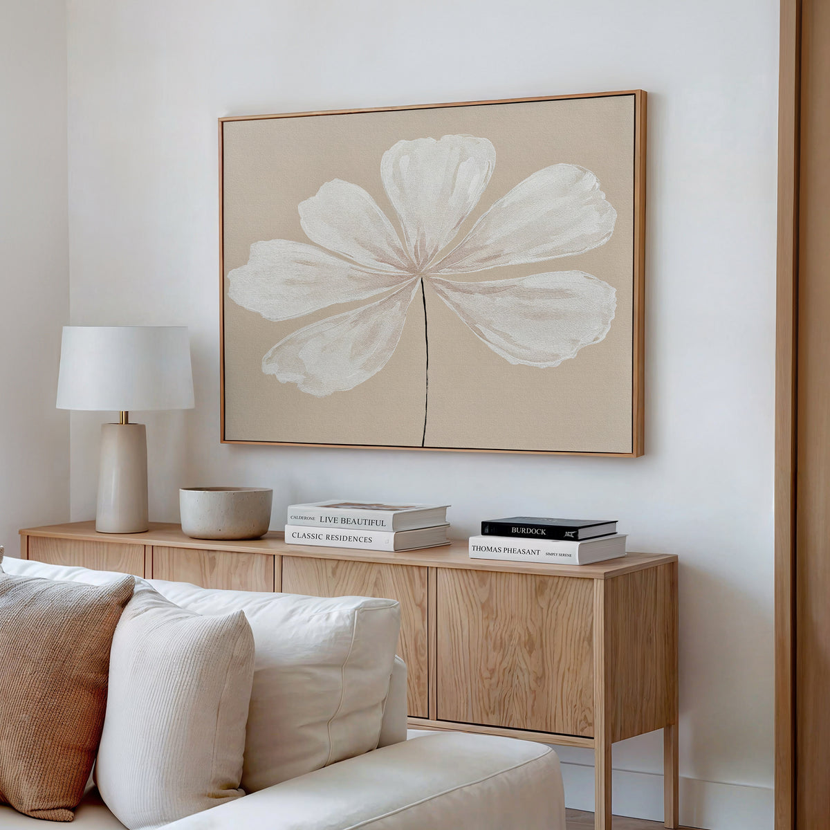 A minimalist living room corner features a light wood sideboard, white lamp, books, and decor. Above it hangs the Ivory Bloom Canvas Art - Horizontal, bringing elegant neutral style. Light sofa cushions are visible in the foreground.