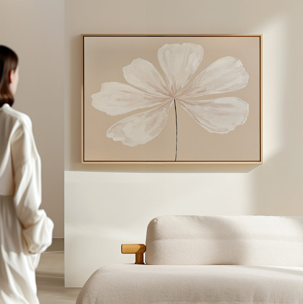 A woman in a white outfit admires the Ivory Bloom Canvas Art - Horizontal, featuring a single white flower on a thin stem, displayed above a beige sofa in a minimalist room with light, neutral décor.
