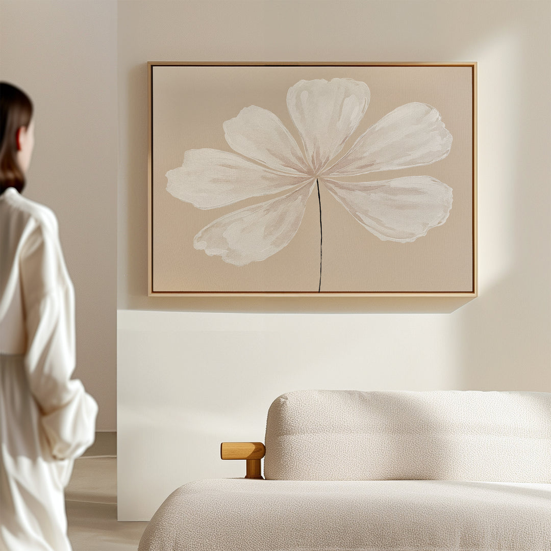 A woman in a white outfit admires the Ivory Bloom Canvas Art - Horizontal, featuring a single white flower on a thin stem, displayed above a beige sofa in a minimalist room with light, neutral décor.